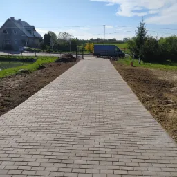 Nowa, długa ścieżka z szarej kostki brukowej prowadząca do metalowej bramy, obok staw i dom z szarą elewacją, błękitne niebo z chmurami.