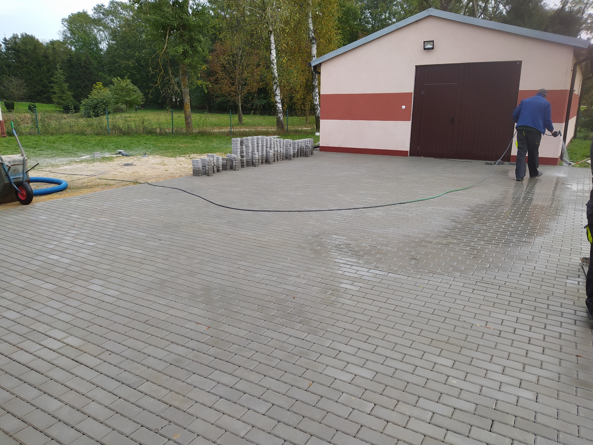 Świeżo ułożona szara kostka brukowa przed budynkiem gospodarczym z brązowymi drzwiami, pracownik czyści powierzchnię wodą pod ciśnieniem, na skraju widoczne ułożone kostki i taczka.