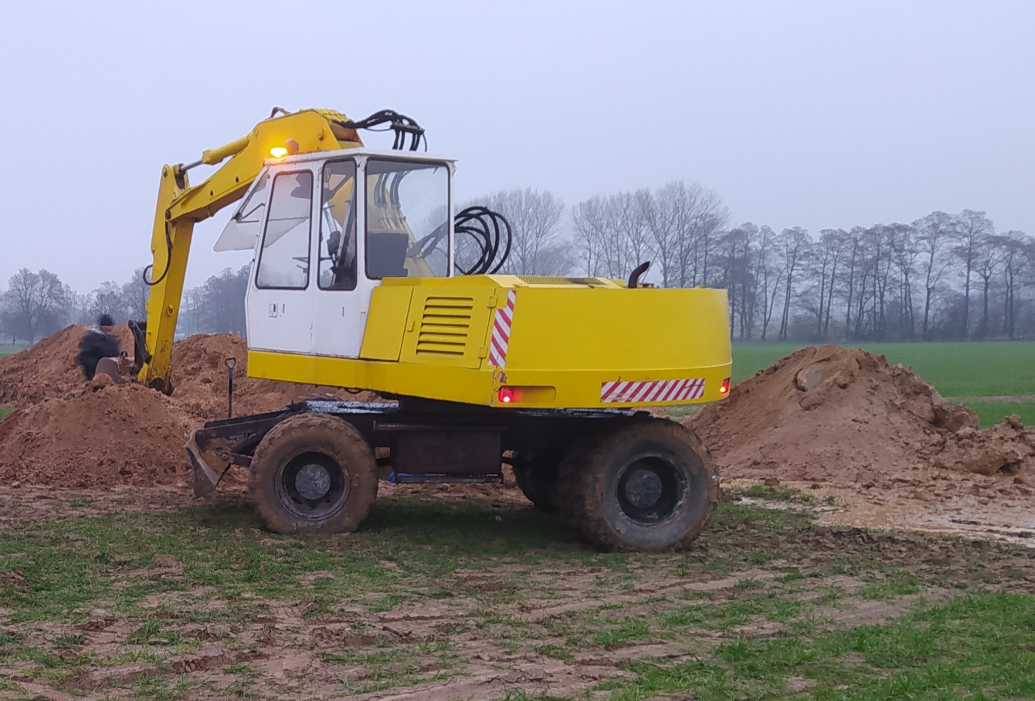 Żółto-biała koparka kołowa na polu, z pracownikiem w tle kopiącym w ziemi, widoczne pryzmy ziemi i drzewa w oddali, pochmurny dzień.