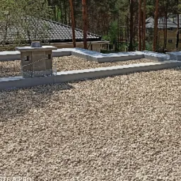 Płaski dach pokryty drobnym, jasnym żwirem, z niskim kominem z szarej cegły i betonowymi obramowaniami, w tle drzewa iglaste i budynek mieszkalny.