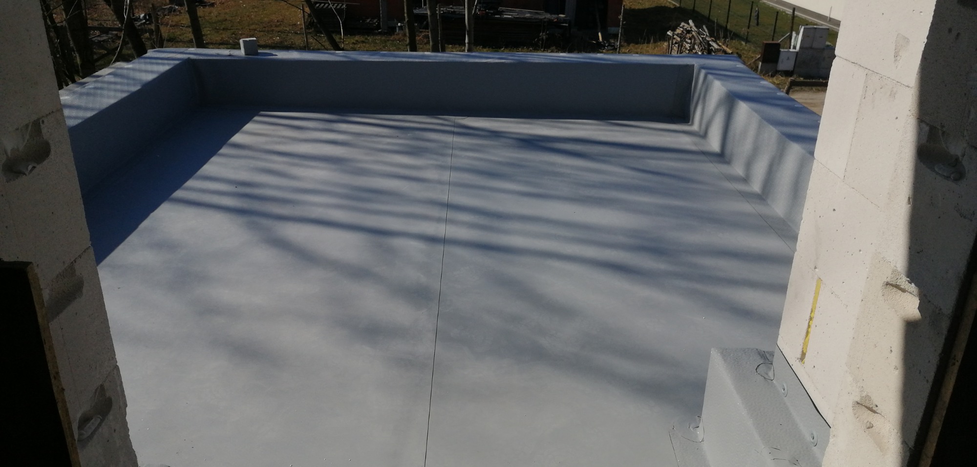 Płaski dach pokryty szarą, jednolitą powłoką hydroizolacyjną, widok z góry, cień drzew na powierzchni, ściany w surowym stanie.