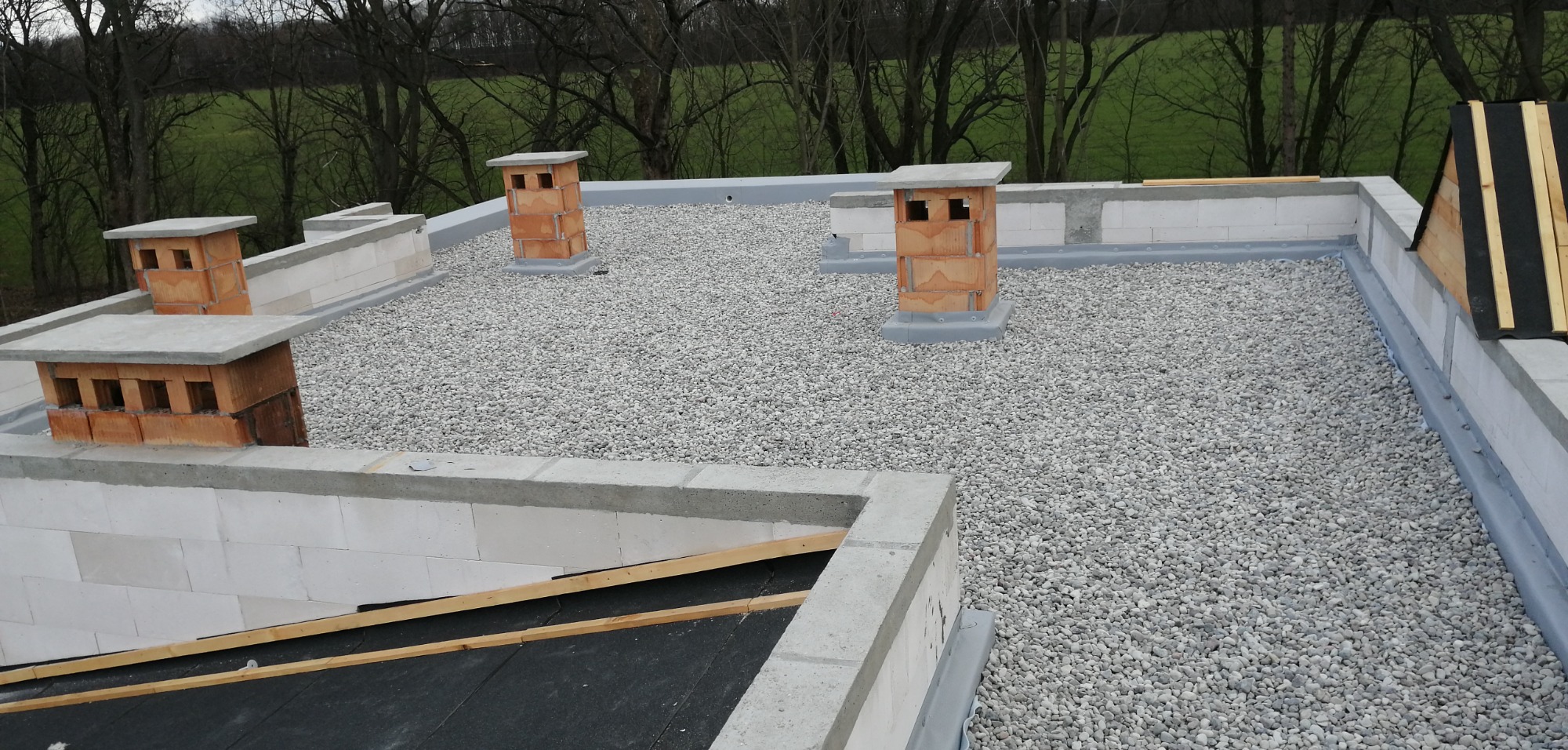 Widok z góry na płaski dach pokryty drobnym żwirem w Ząbkach, z widocznymi kominami z cegły klinkierowej i fragmentami konstrukcji obłożonej bloczkami betonowymi.