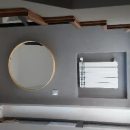 Szara ściana z okrągłym lustrem w złotej ramie, grzejnikiem panelowym i drewnianymi elementami dekoracyjnymi pod sufitem, oświetlona światłem dziennym.