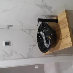 Czarna umywalka nablatowa z marmurowym wzorem i czarną armaturą na drewnianym blacie, na tle białych marmurowych płytek ściennych i drewnianej podłogi w jodełkę.