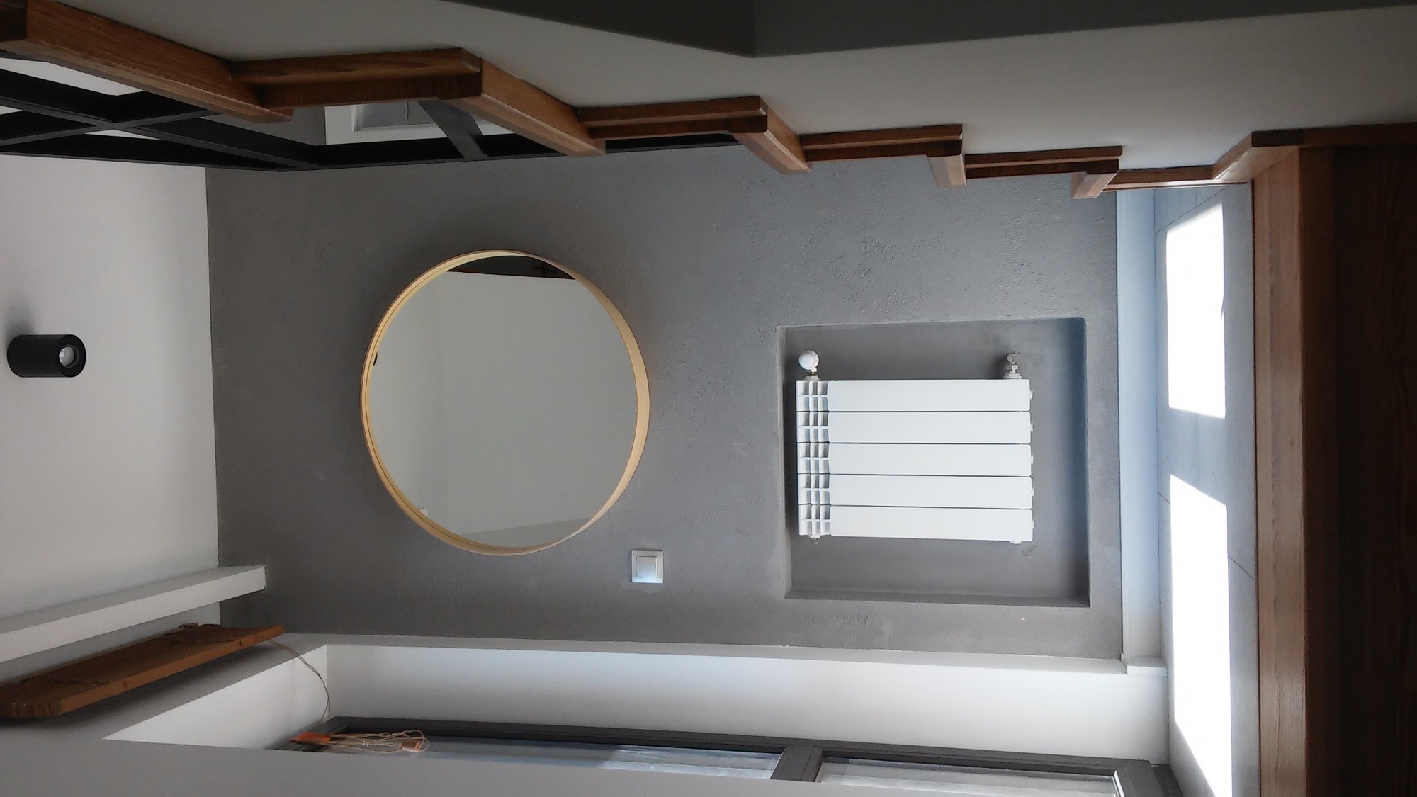 Szara ściana z okrągłym lustrem w złotej ramie, grzejnikiem panelowym i drewnianymi elementami dekoracyjnymi pod sufitem, oświetlona światłem dziennym.