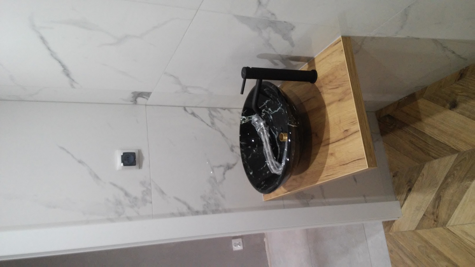 Czarna umywalka nablatowa z marmurowym wzorem i czarną armaturą na drewnianym blacie, na tle białych marmurowych płytek ściennych i drewnianej podłogi w jodełkę.