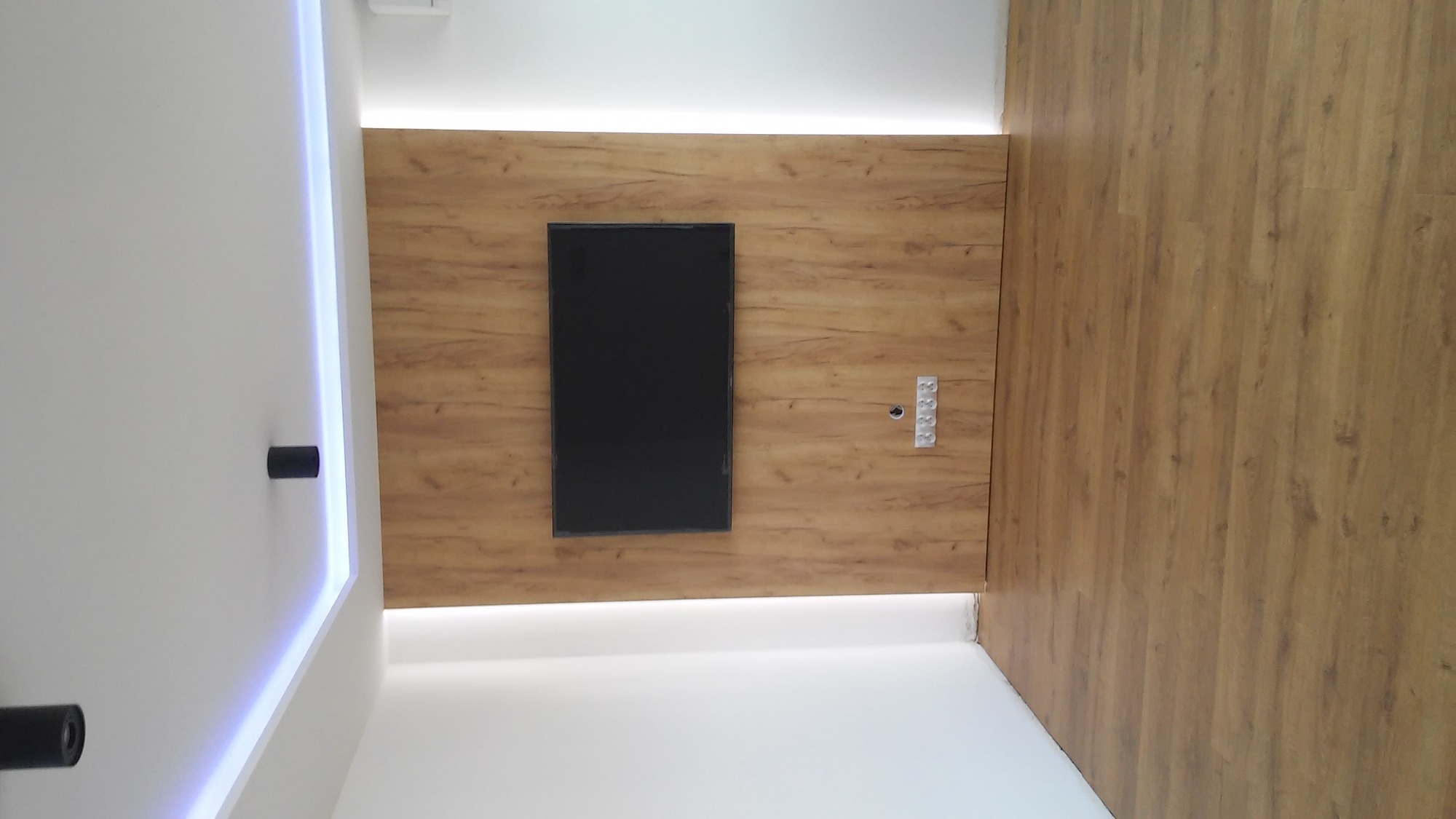 Nowoczesny salon z drewnianą ścianą i podłogą, podświetlany sufit z czarnymi reflektorami, centralnie umieszczony czarny telewizor.