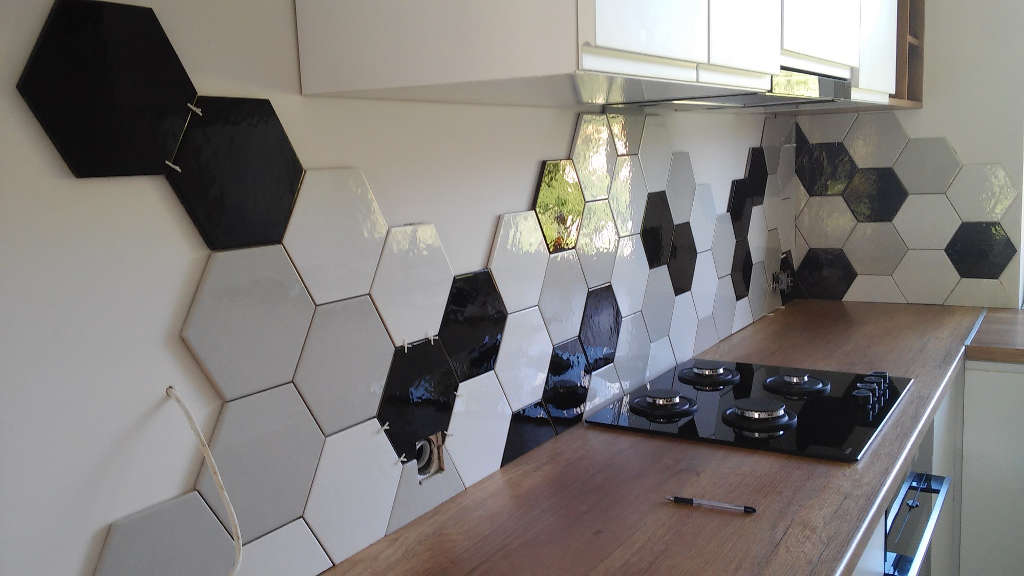 Układanie heksagonalnych płytek w kolorach czerni, bieli i szarości na ścianie nad blatem kuchennym, widoczne krzyżyki dystansowe i otwór na gniazdko elektryczne.