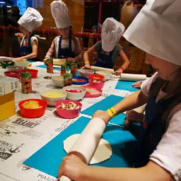 Dzieci w czapkach kucharskich wałkują ciasto na pizzę podczas warsztatów kulinarnych, na stole kolorowe miseczki z dodatkami i soki Tymbark.