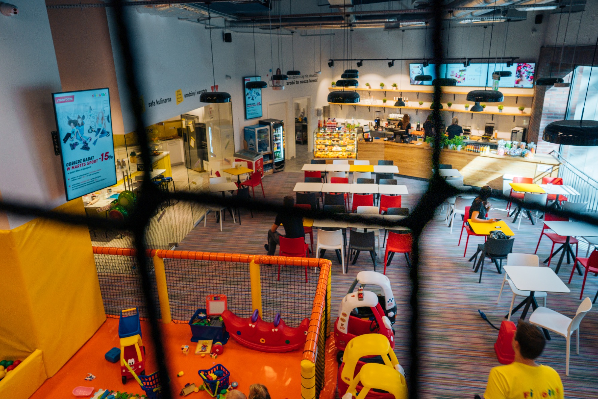 Przestronne wnętrze kawiarni z wydzieloną strefą zabaw dla dzieci, widok z góry przez siatkę zabezpieczającą, w tle lada z ciastami i ekspres do kawy.