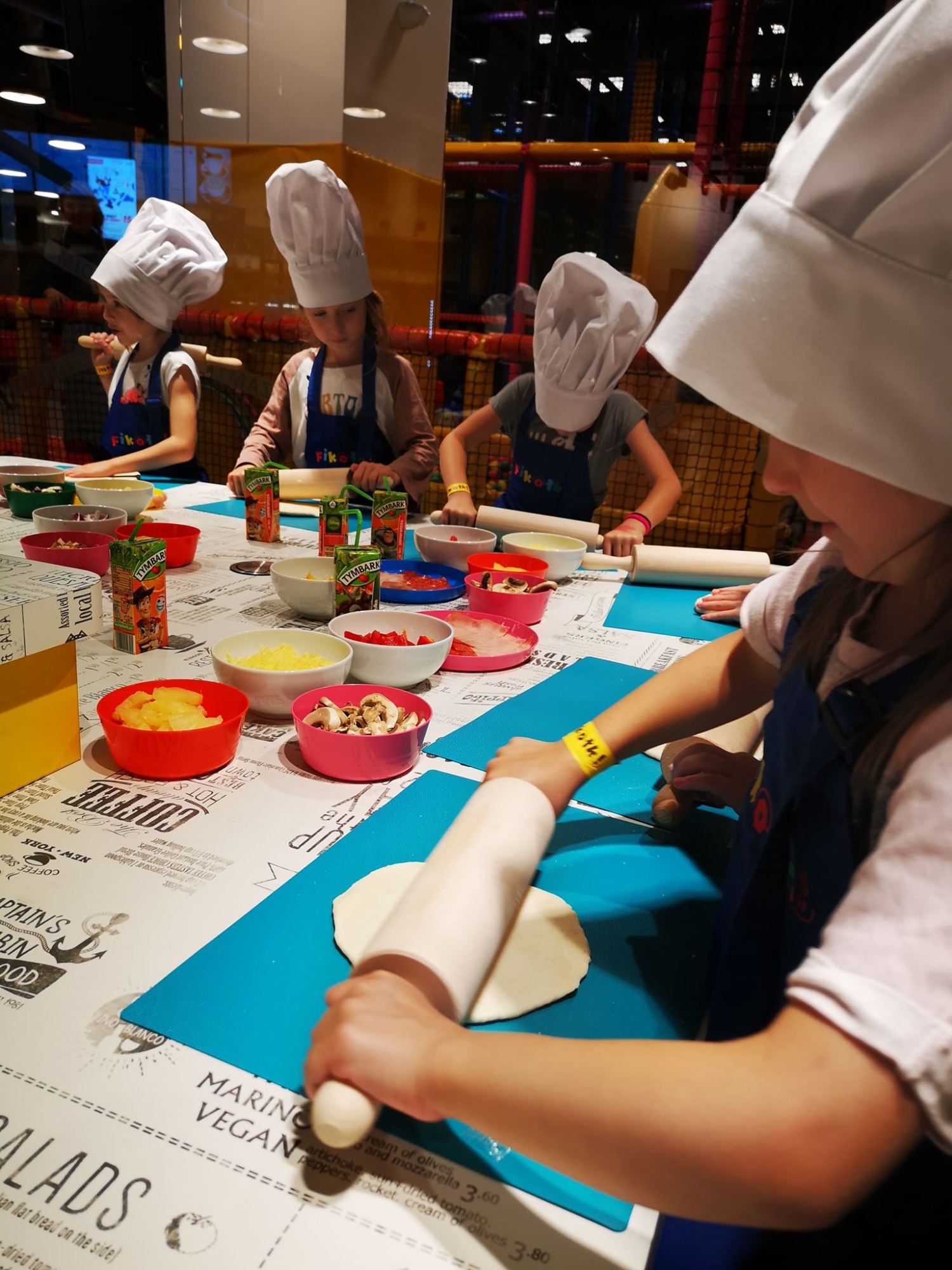 Dzieci w czapkach kucharskich wałkują ciasto na pizzę podczas warsztatów kulinarnych, na stole kolorowe miseczki z dodatkami i soki Tymbark.