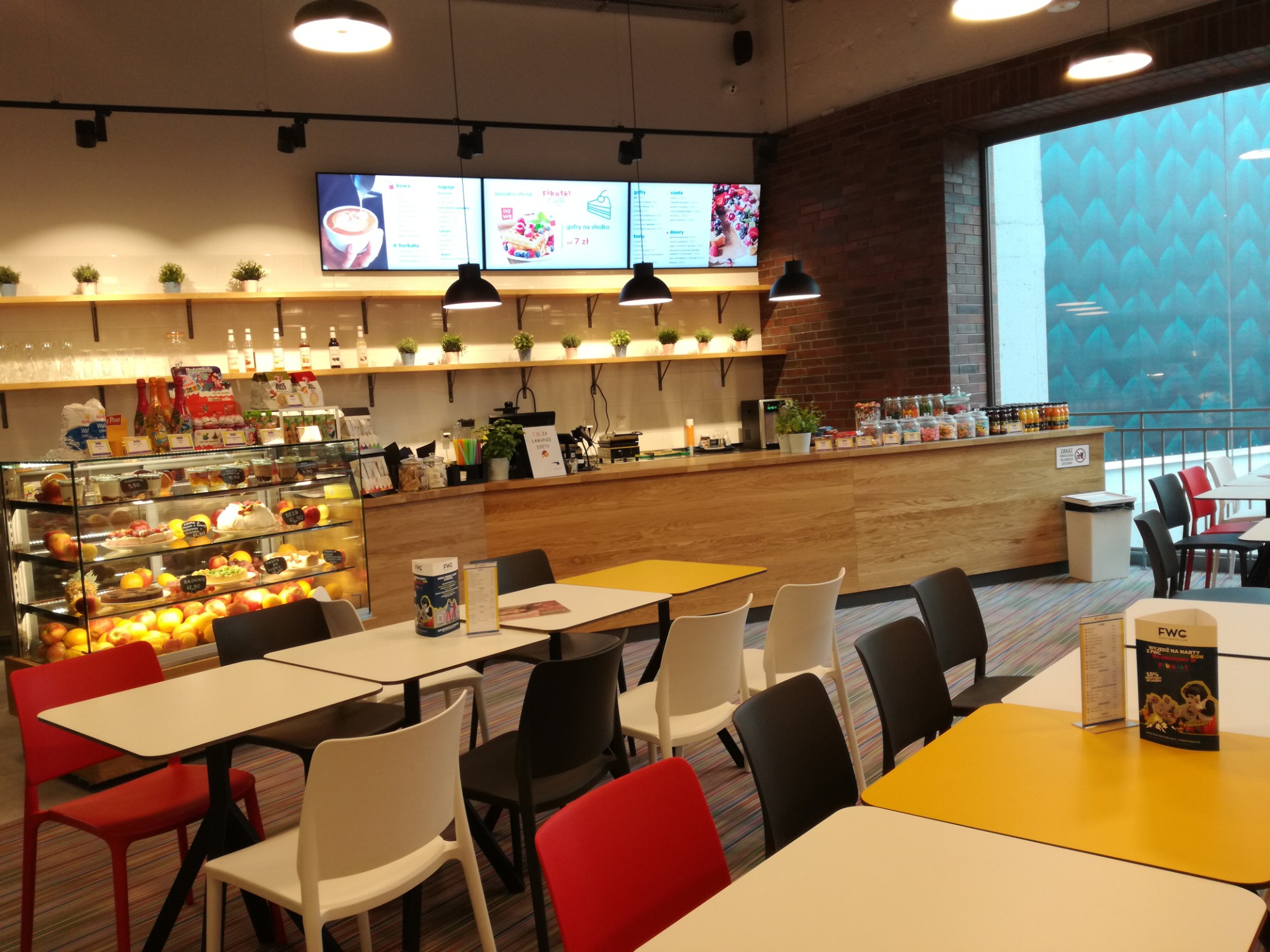 Wnętrze kawiarni z przeszkloną witryną pełną ciast i owoców, ekspozycją napojów na półkach, menu na ekranach, drewnianym blatem i stolikami z krzesłami w kolorach białym, czarnym, czerwonym i żółtym.