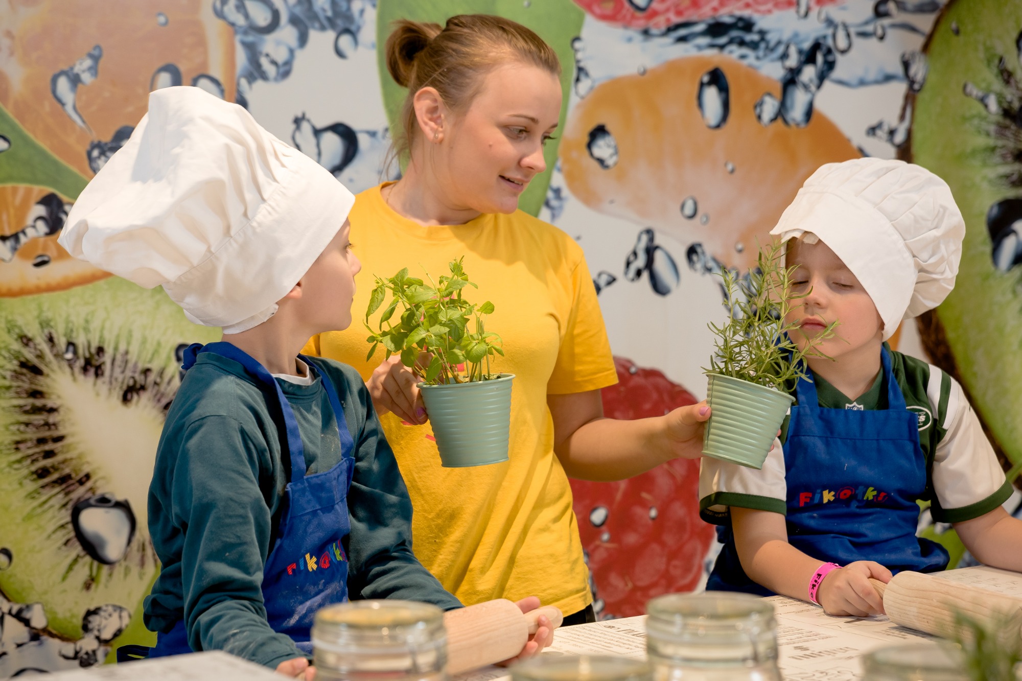Dzieci w czapkach kucharskich i fartuchach na warsztatach kulinarnych, wąchają zioła, a instruktorka trzyma doniczkę z bazylią, w tle dekoracja z owocami.