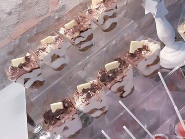 Deser tiramisu w przezroczystych, kwadratowych pojemniczkach ułożonych na schodkowej ekspozycji, udekorowany kawałkiem białej czekolady.