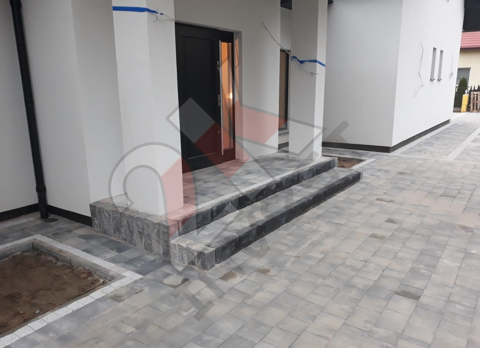 Nowoczesne wejście do domu z kostki brukowej w odcieniach szarości, schody z obrzeżami z ciemniejszej kostki, widoczne ślady po deszczu na powierzchni.