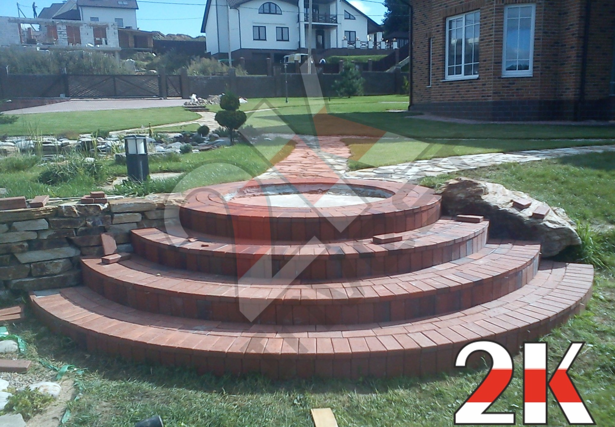 Okrągłe schody z czerwonej cegły prowadzące do małego basenu lub ogniska, otoczone trawnikiem i elementami ogrodu, w tle dom z cegły i ogród.