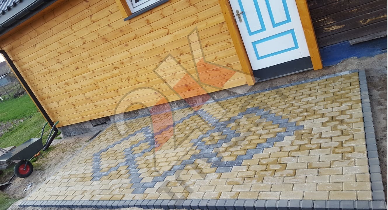 Ułożona kostka brukowa w odcieniach żółci i szarości tworząca wzór przed drewnianym domkiem z niebiesko-białymi drzwiami. W tle widoczny wózek budowlany.