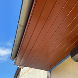 Widok z dołu na pomalowaną na brązowo podbitkę dachową z widoczną rynną i fragmentem elewacji zabezpieczonej niebieską taśmą malarską.