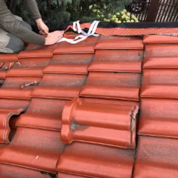 Dekarz mocuje membranę dachową do dachówki ceramicznej, widoczne białe taśmy mocujące, w tle iglaki i fragment ogrodzenia.