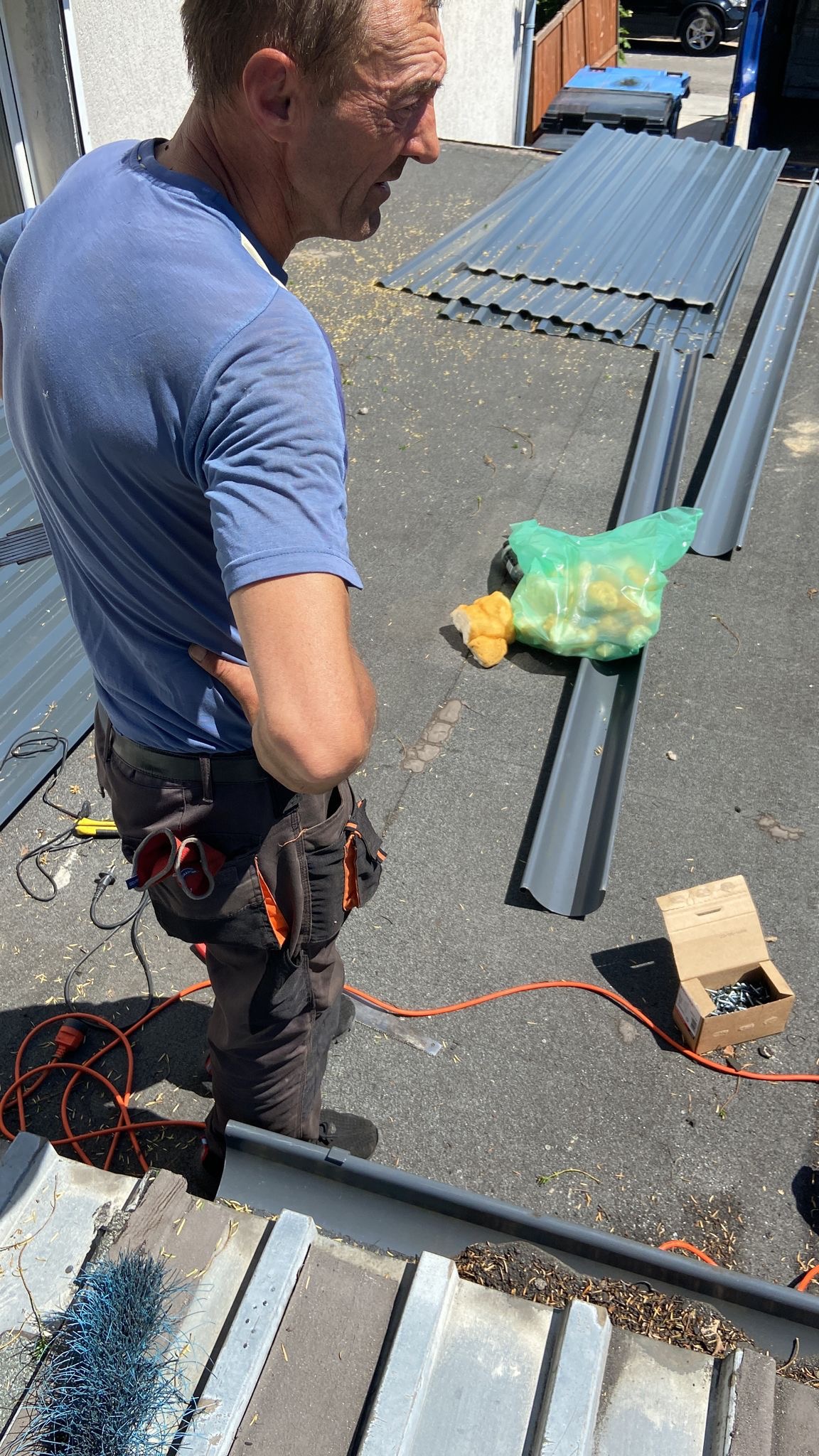 Dekarz na dachu przygotowuje się do montażu blachy trapezowej i rynien, w tle zapas blachy, narzędzia w kieszeniach, worek z żółtymi elementami mocującymi.