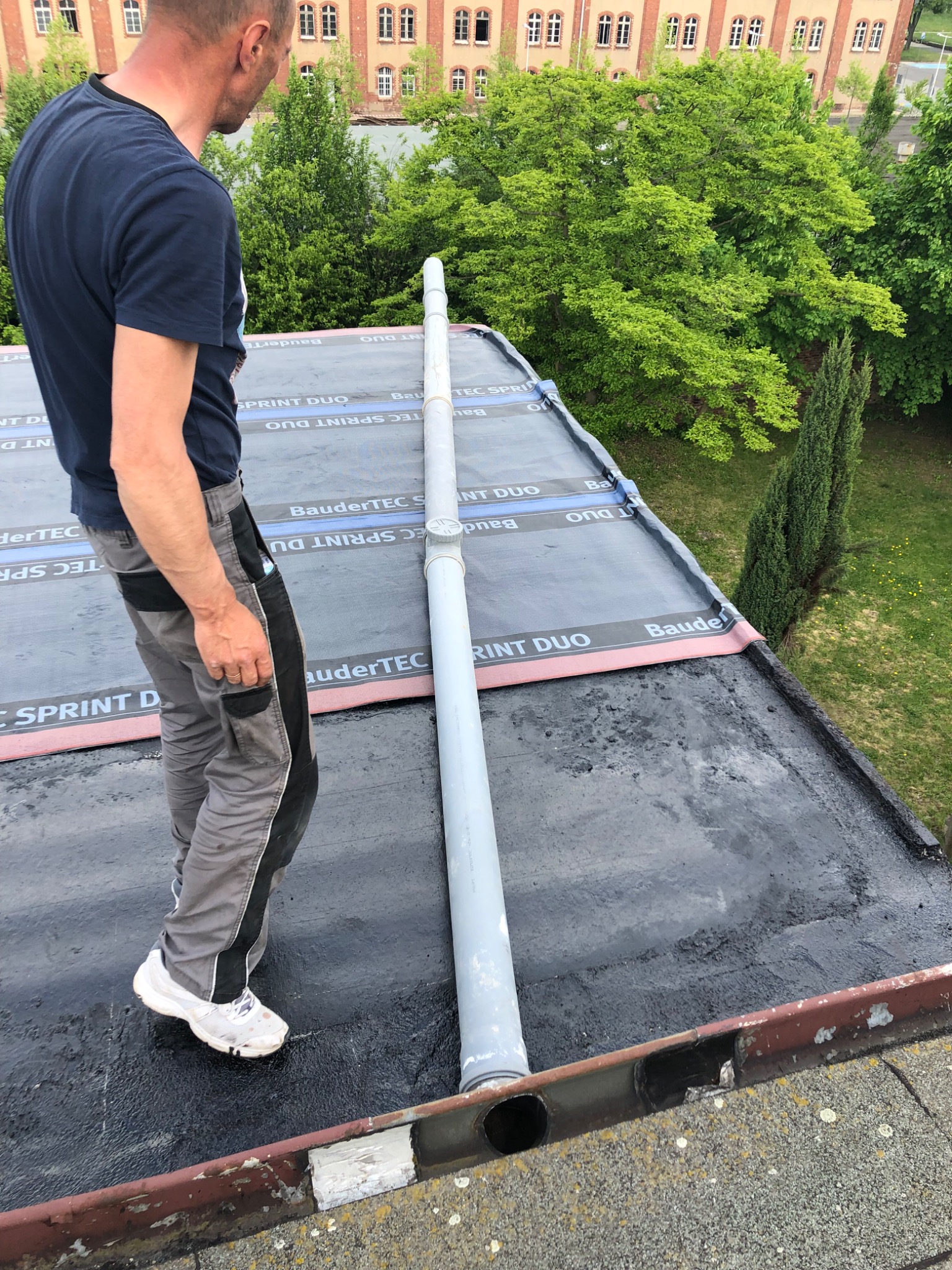 Pracownik w szarych spodniach roboczych i białych butach stoi na czarnym, płaskim dachu pokrytym papą z widocznym systemem odprowadzania wody deszczowej, na tle zielonych drzew i budynku...