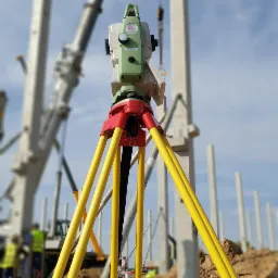 Zielono-biały teodolit Leica na żółtym statywie, ustawiony na placu budowy z widocznymi słupami konstrukcyjnymi i dźwigiem w tle, z ziemią w pierwszym planie.