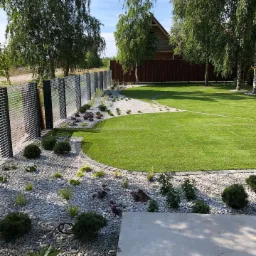 Starannie zaprojektowany ogród z równo przystrzyżonym trawnikiem, białymi kamieniami, ozdobnymi krzewami i nowoczesnym ogrodzeniem panelowym na tle domu i drzew.