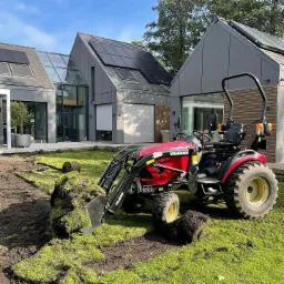 Czerwony traktor Yanmar 424 z łychą wypełnioną rolkami trawy na tle nowoczesnego domu z panelami słonecznymi w Sławoszynie, przygotowania do rozkładania trawy.