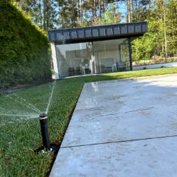 Automatyczny system nawadniania trawnika z rolki, zraszacze w akcji. Nowoczesny dom z dużymi oknami w tle, świeżo założony trawnik i jasne płytki chodnikowe.