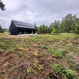 A-Z Ogród Damian Mroch - Nowoczesny dom z panelami słonecznymi w trakcie prac ogrodowych. Widoczny przygotowany teren pod ogród i osoba pracująca w oddali. Pochmurne niebo.