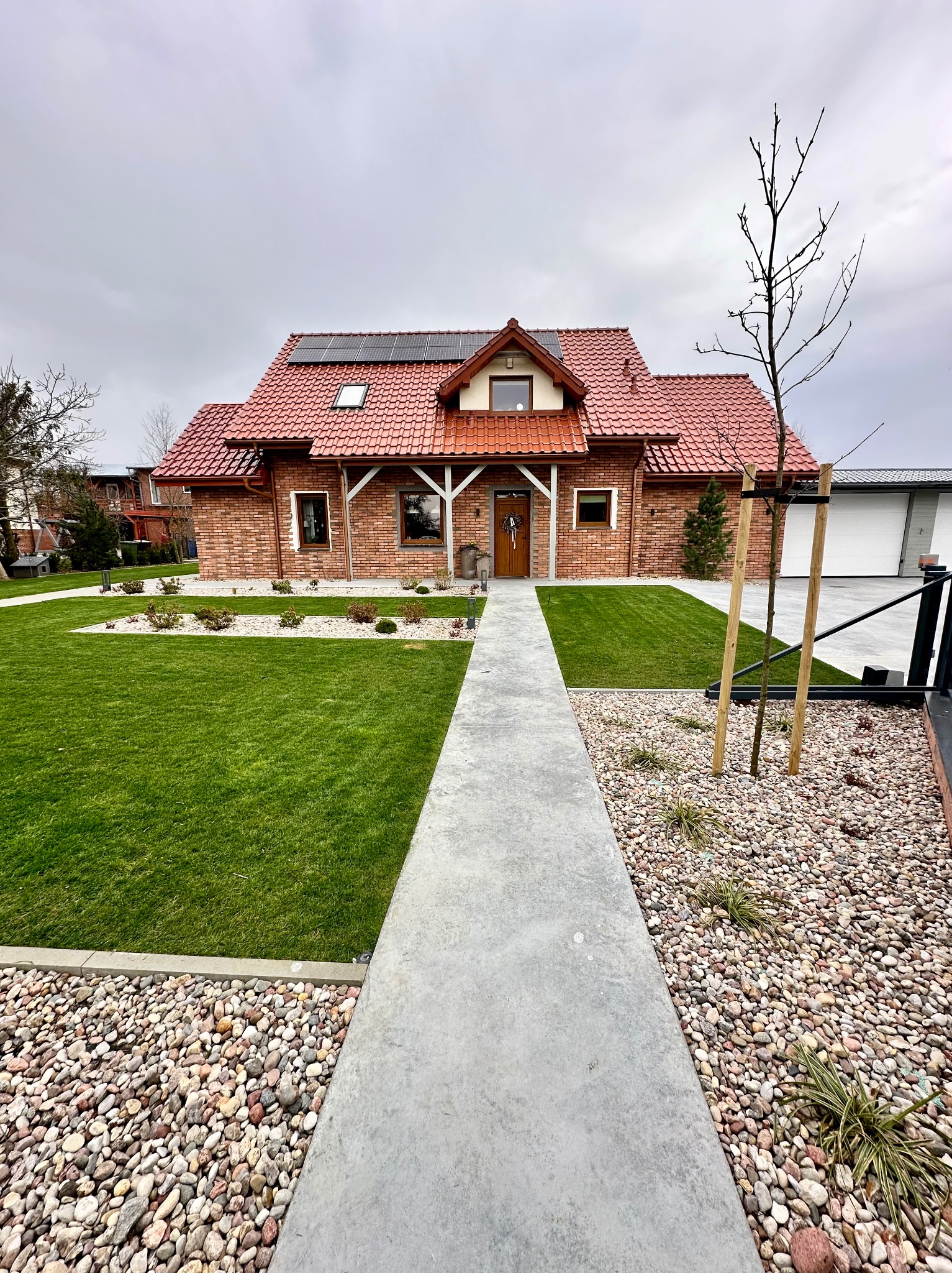Bryła domu z czerwonej cegły z zadbanym trawnikiem i ścieżką z kamieniami. Na dachu panele słoneczne, a obok domu garaż z bramą.