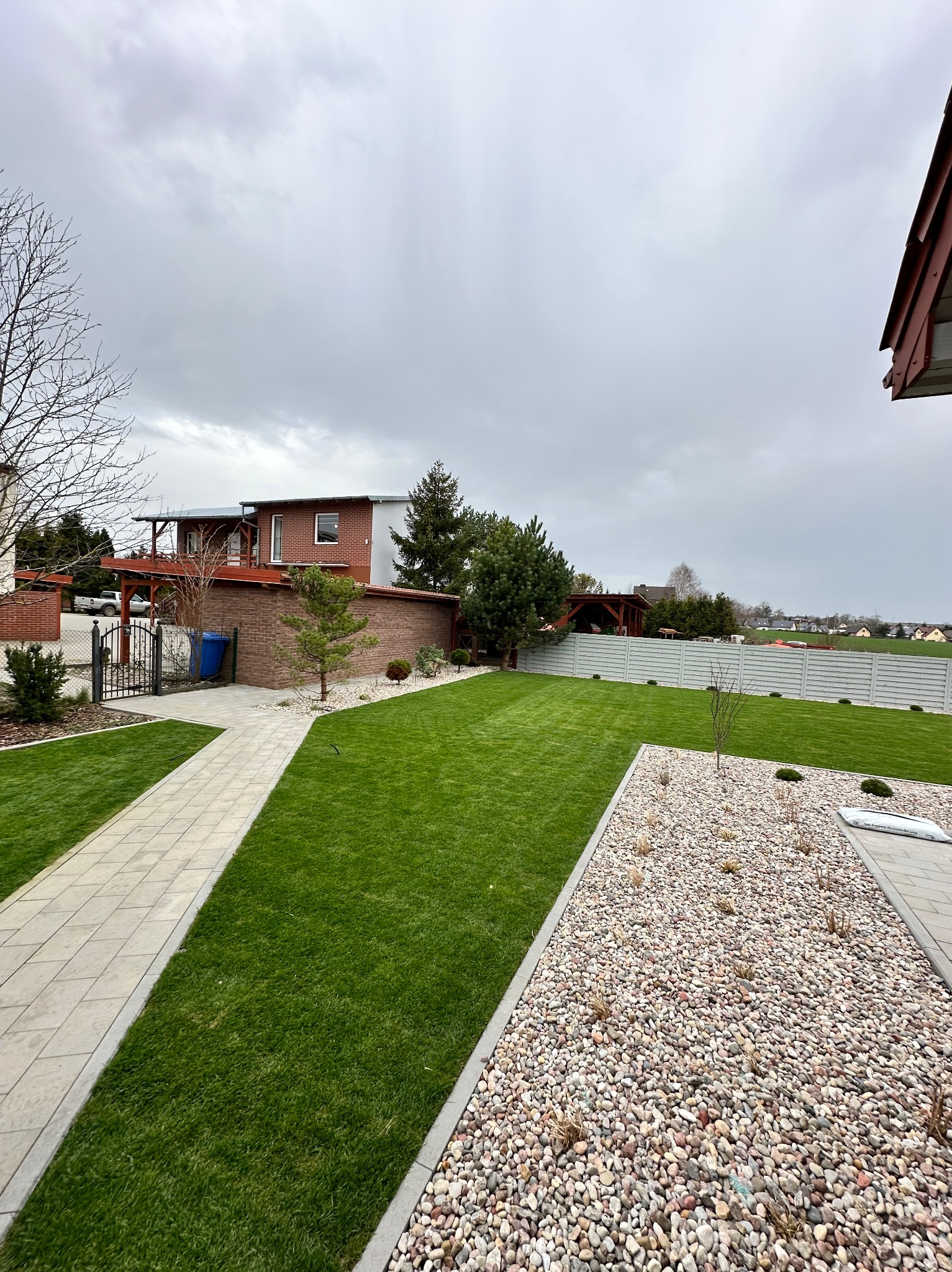 Zadbany ogród z równo przystrzyżonym trawnikiem, ścieżką z płyt i rabatą z kamieni. W tle dom z cegły i drewniana altana, całość pod pochmurnym niebem.