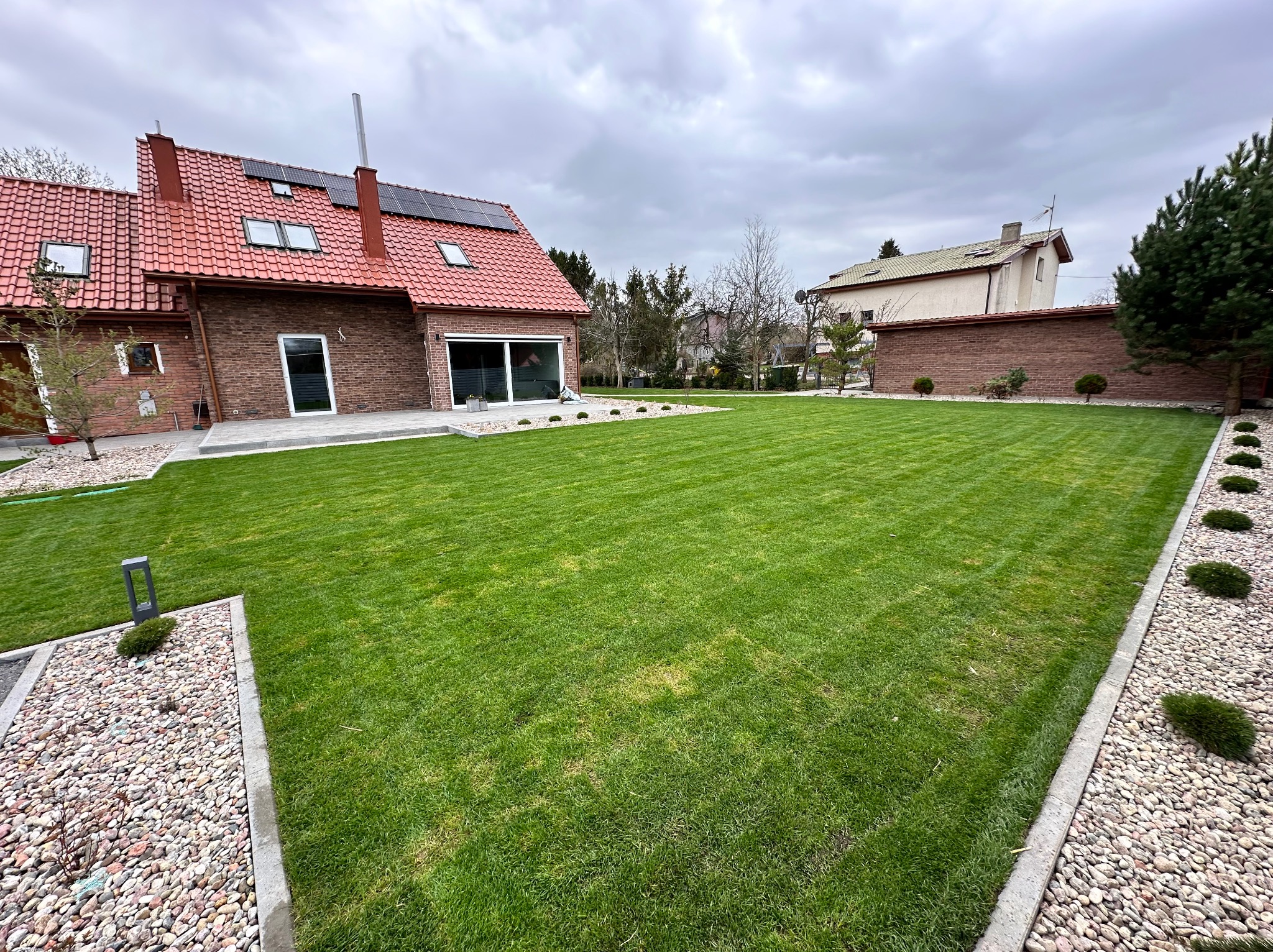 Perfekcyjnie skoszony trawnik przed nowoczesnym domem z czerwoną dachówką i panelami słonecznymi. Widoczny kamienny chodnik i ozdobne nasadzenia.