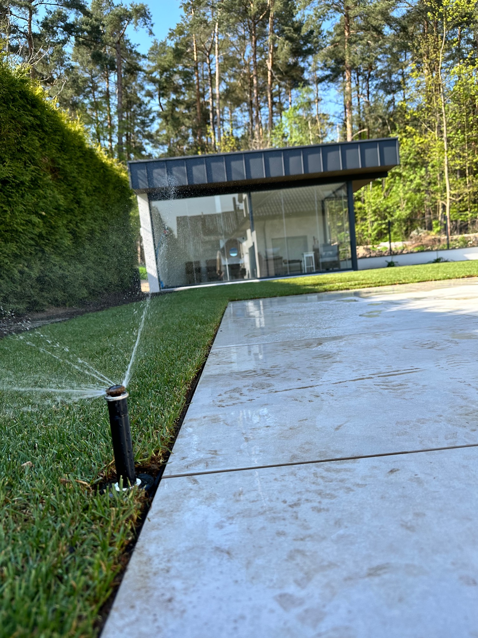 Automatyczny system nawadniania trawnika z rolki, zraszacze w akcji. Nowoczesny dom z dużymi oknami w tle, świeżo założony trawnik i jasne płytki chodnikowe.