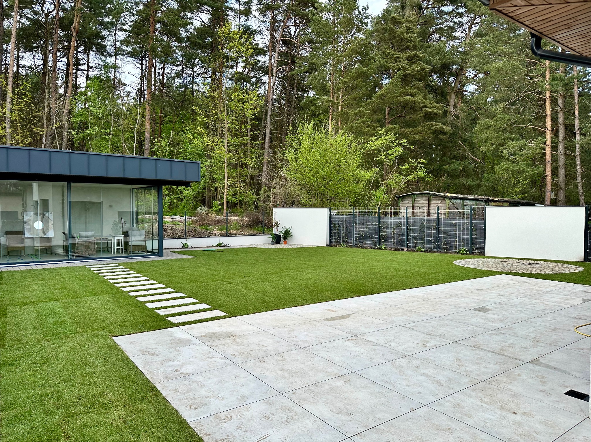 Nowoczesny ogród z równo przystrzyżonym trawnikiem, ścieżką z płyt i tarasem z jasnych płytek. W tle zabudowa domu z dużymi oknami i zielone drzewa.