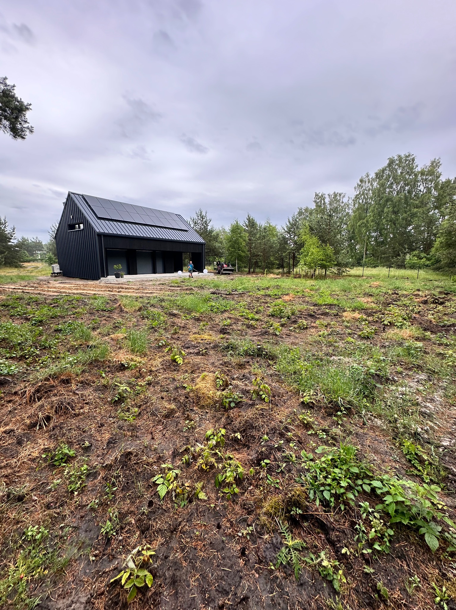 Nowoczesny dom z panelami słonecznymi w trakcie prac ogrodowych. Widoczny przygotowany teren pod ogród i osoba pracująca w oddali. Pochmurne niebo.
