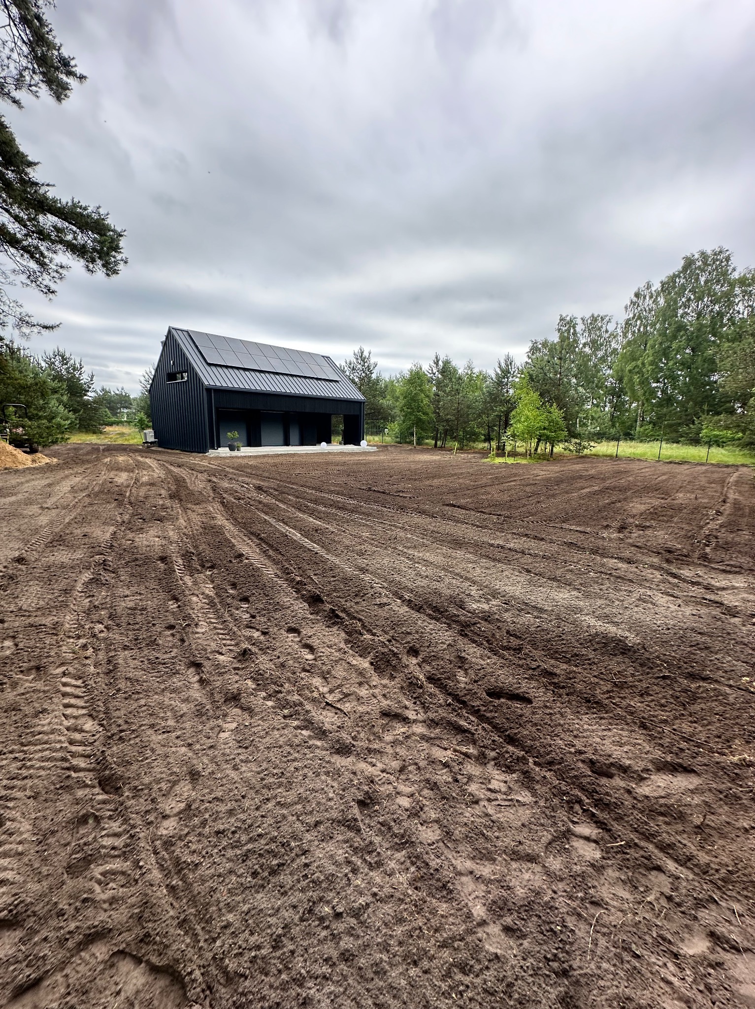 Przygotowany teren pod ogród z widokiem na nowoczesny dom z panelami słonecznymi. Ślady opon na ziemi sugerują niedawne prace ziemne.