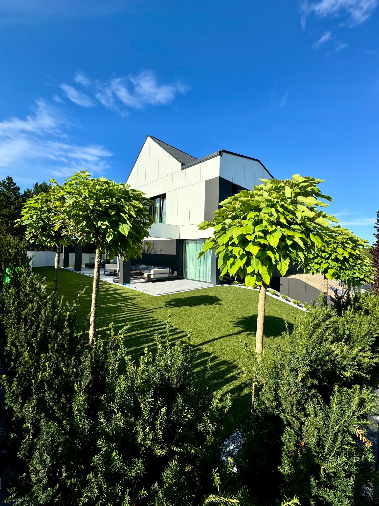 Nowoczesny dom z zadbanym ogrodem: równo przystrzyżona trawa, kuliste drzewa i ozdobne krzewy. Słoneczny dzień, błękitne niebo z delikatnymi chmurami.