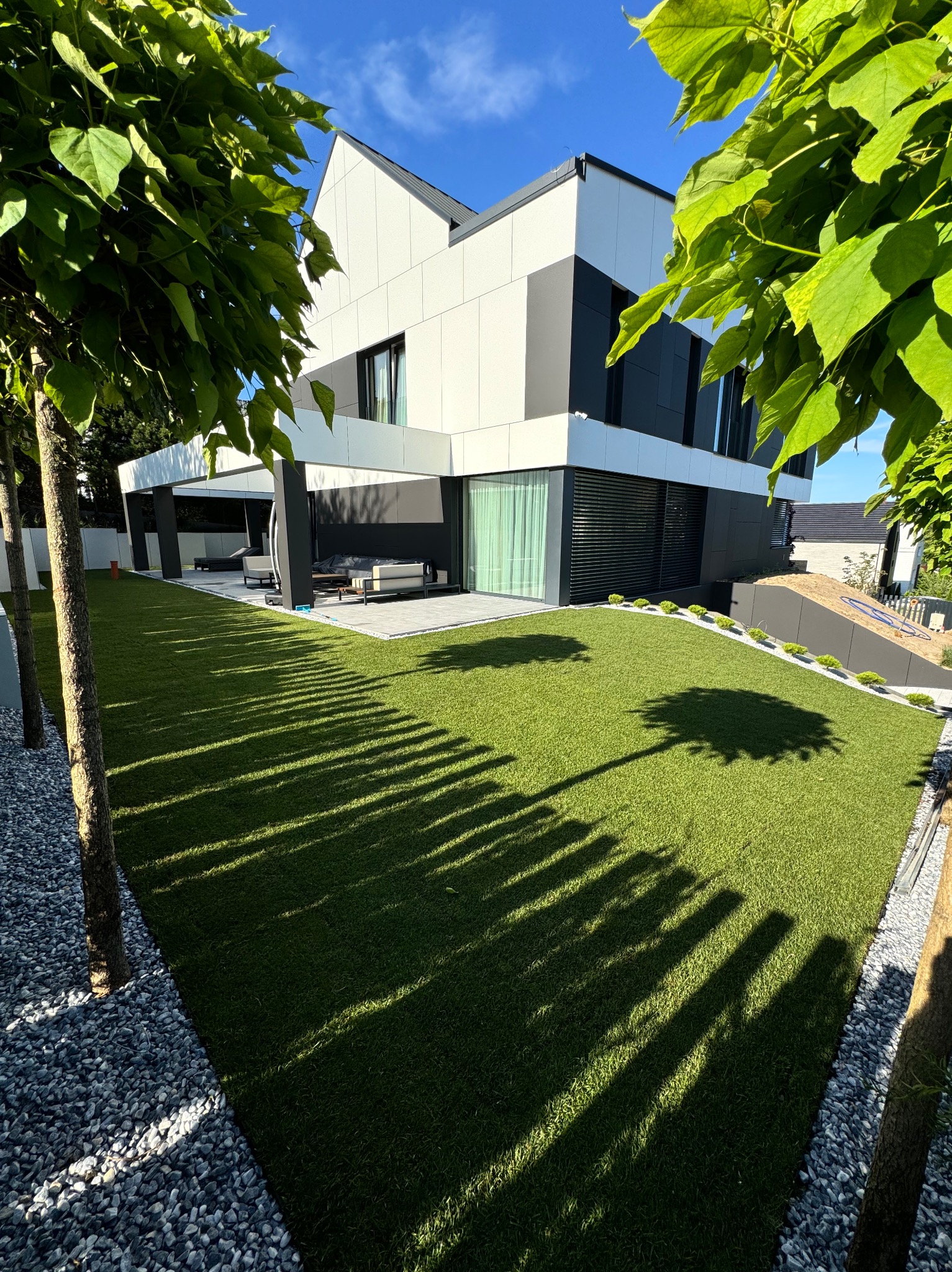 Nowoczesny ogród z równo przystrzyżonym trawnikiem, drzewami wzdłuż ścieżki z kamieni i minimalistycznym domem w tle, oświetlony jasnym słońcem.