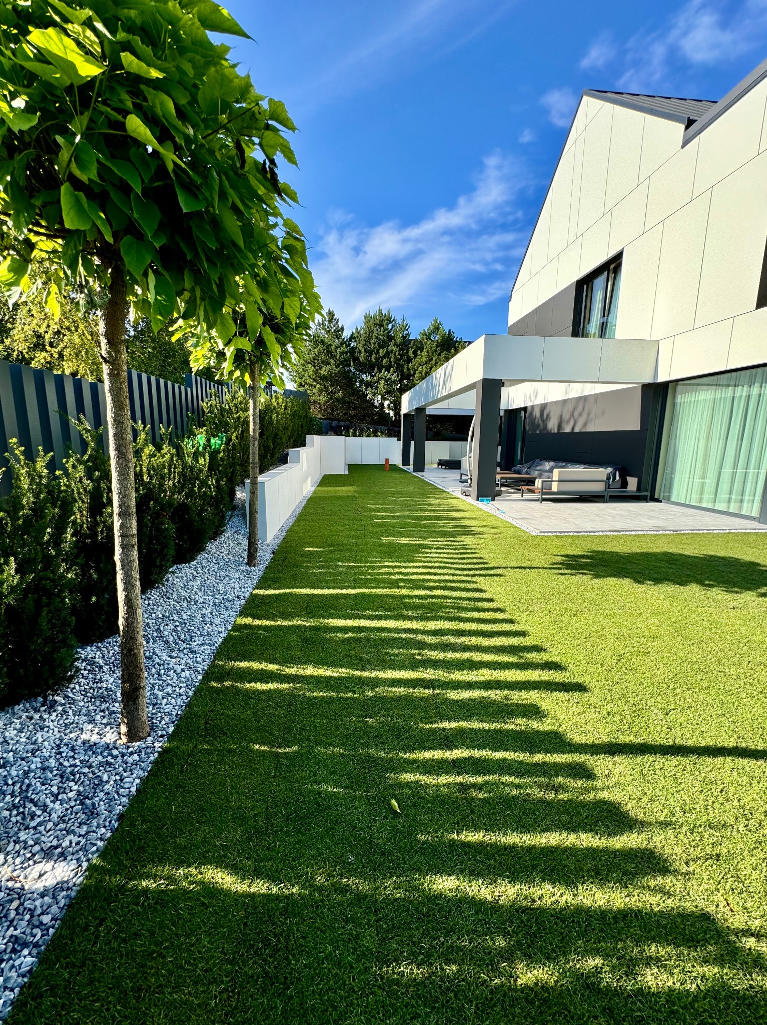 Nowoczesny ogród z równo przystrzyżoną trawą i drzewami kulistymi, obok kamienny żwir. W tle dom z tarasem i błękitne niebo.
