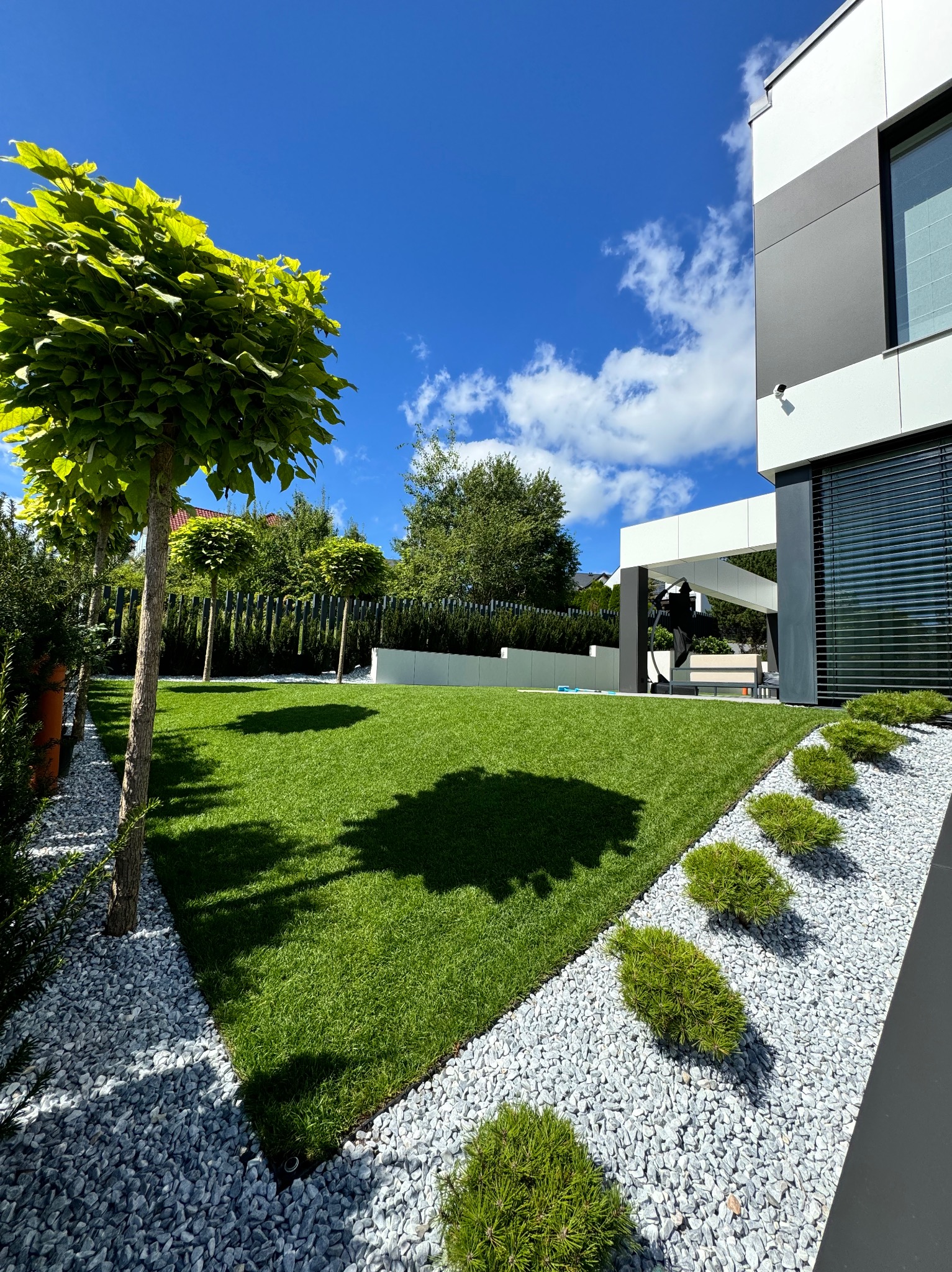 Nowoczesny ogród z równo przystrzyżonym trawnikiem, ozdobnymi drzewami i białymi kamieniami. Architektura budynku w tle. Słoneczny dzień, błękitne niebo.