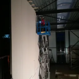 Montaż płyt karton-gips na ścianie hali przemysłowej z wykorzystaniem podnośnika koszowego przez pracownika w czerwonej bluzie, widoczne drewniane elementy konstrukcyjne pod sufitem.