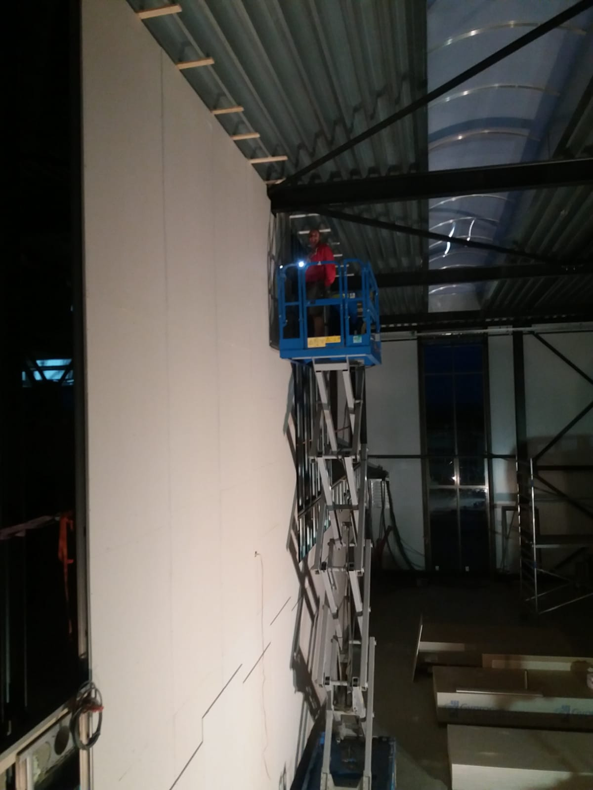 Montaż płyt karton-gips na ścianie hali przemysłowej z wykorzystaniem podnośnika koszowego przez pracownika w czerwonej bluzie, widoczne drewniane elementy konstrukcyjne pod sufitem.