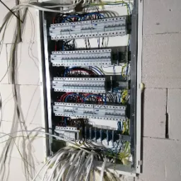 Rozdzielnica elektryczna w trakcie montażu w surowym wnętrzu, widoczne liczne przewody, listwy zaciskowe i elementy zabezpieczające, ściana z betonu komórkowego.