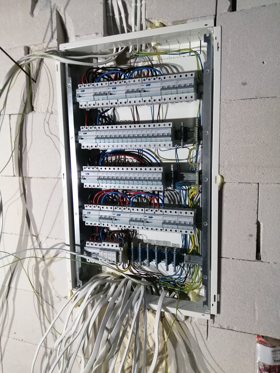 Rozdzielnica elektryczna w trakcie montażu w surowym wnętrzu, widoczne liczne przewody, listwy zaciskowe i elementy zabezpieczające, ściana z betonu komórkowego.