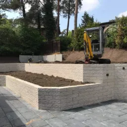 Tarasy ogrodowe z jasnego kamienia, wyrównany teren z mini koparką w tle, świeżo ułożona kostka brukowa na pierwszym planie.