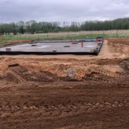 Wylany fundament pod budynek, zabezpieczony folią, z wystającymi rurami instalacyjnymi, otoczony świeżo wyrównanym terenem z widocznymi śladami opon maszyn budowlanych, w tle łąka i las...