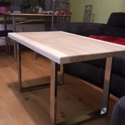 Stolik kawowy z blatem z jasnego drewna i polerowanymi, metalowymi nogami o kwadratowym przekroju, ustawiony na drewnianej podłodze w salonie z ciemną sofą w tle.