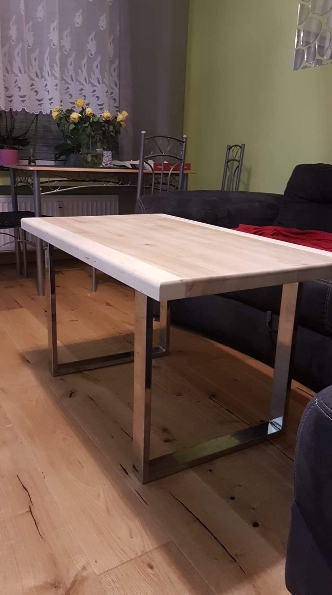 Stolik kawowy z blatem z jasnego drewna i polerowanymi, metalowymi nogami o kwadratowym przekroju, ustawiony na drewnianej podłodze w salonie z ciemną sofą w tle.