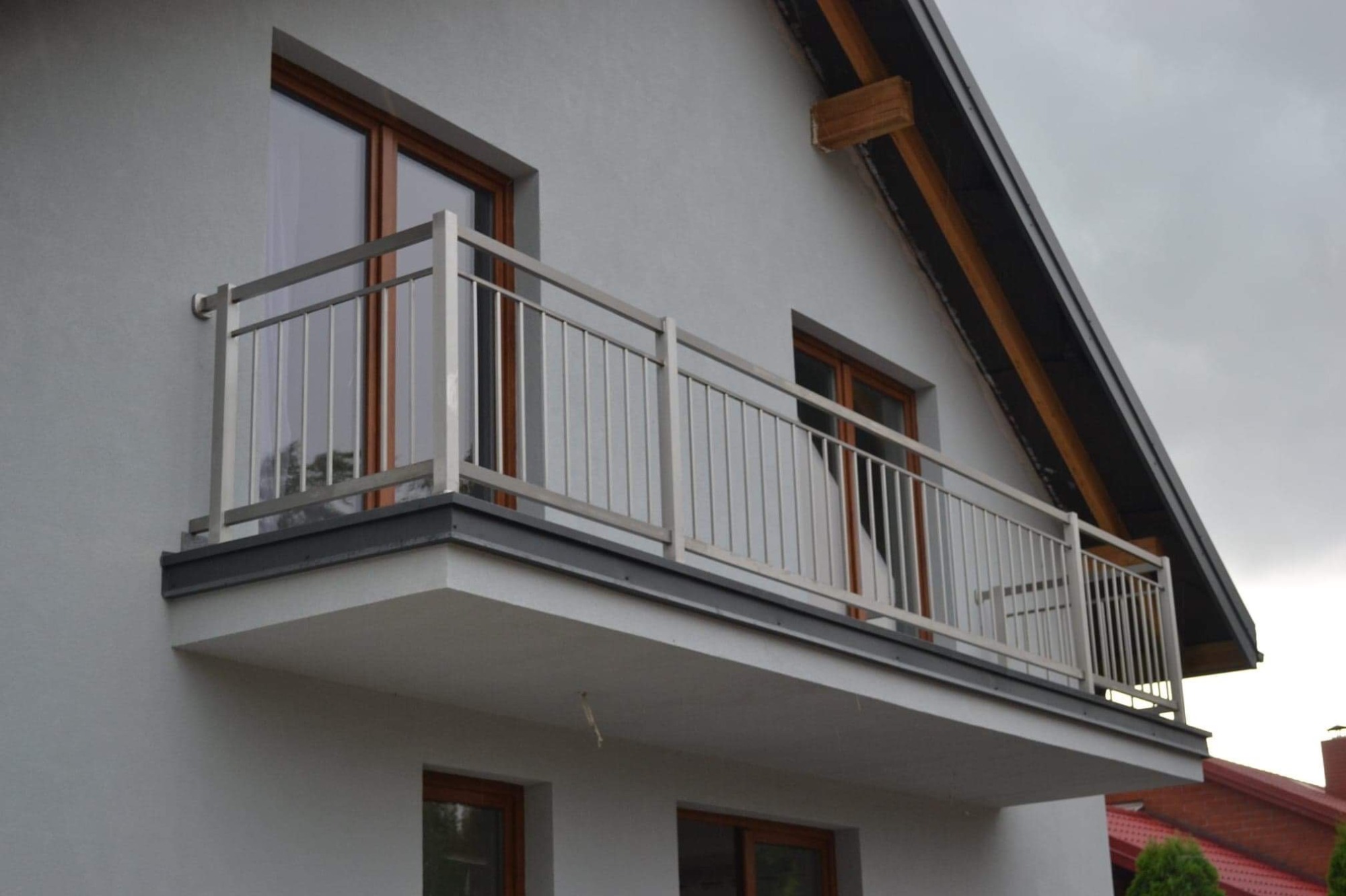 Balkon z nowoczesną, metalową balustradą o minimalistycznym designie, widok z dołu, szara elewacja budynku z drewnianymi ramami okiennymi.